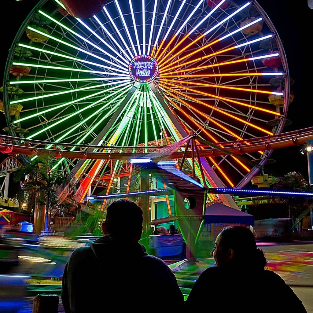 Two visitors look at red, white, and green colors on the Ferris wheel for Cinco de Mayo at the Santa Monica Pier | Photo by @plaztikz0ul