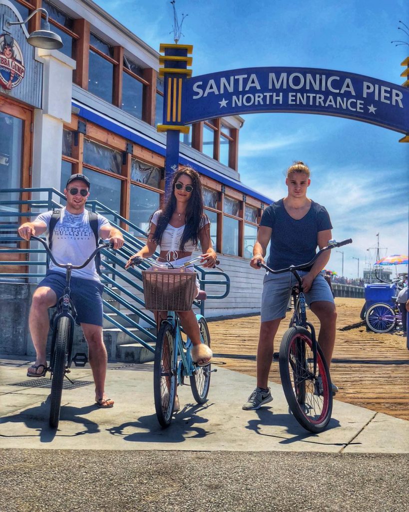 A woman and two men on bikes in front of a blue sign for the Santa Monica Pier
