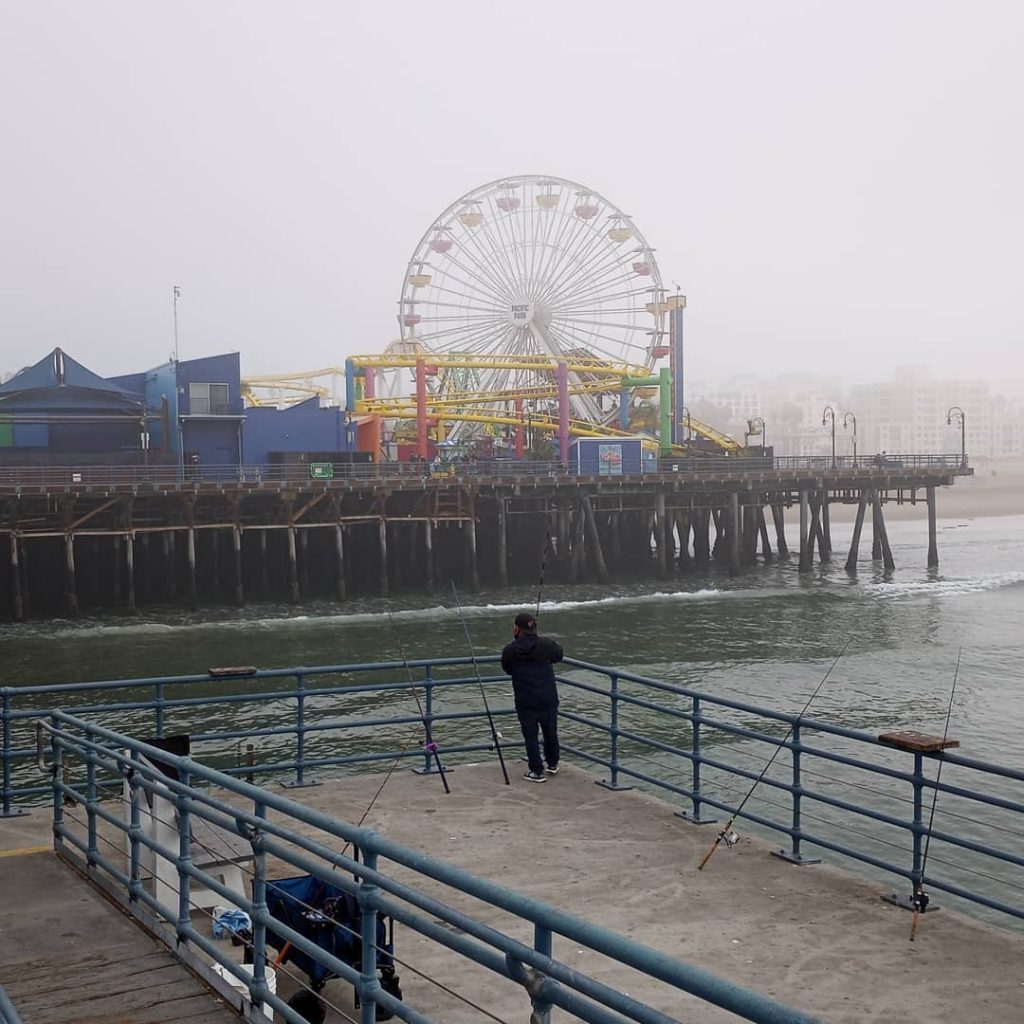 A fisherman on the Santa Monica Pier with the skyline obscured by a dense marine layer - Photo by @daniel_flores333