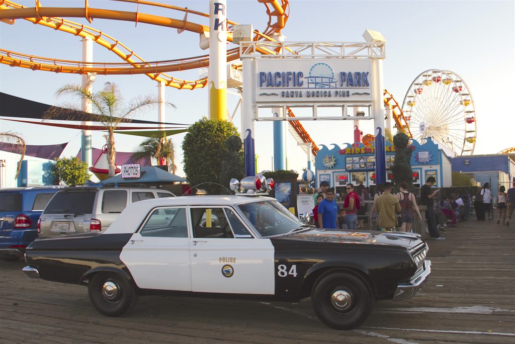 A classic Plymouth police cruiser parked in front of the entrance sign to Pacific Park on the Santa Monica Pier