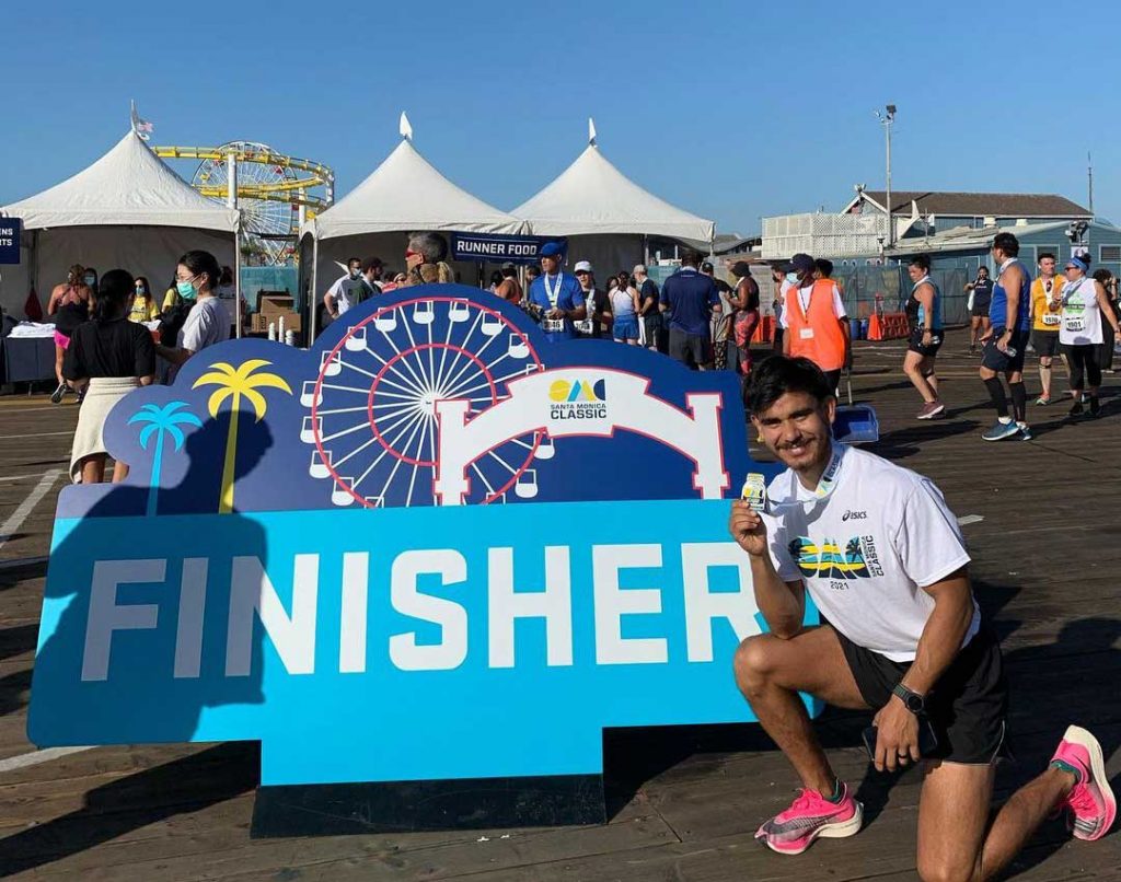 A runner kneels by a bright blue finisher sign on the Santa Monica pier after completing the Santa Monica Classic | Photo by @_juaneezy_