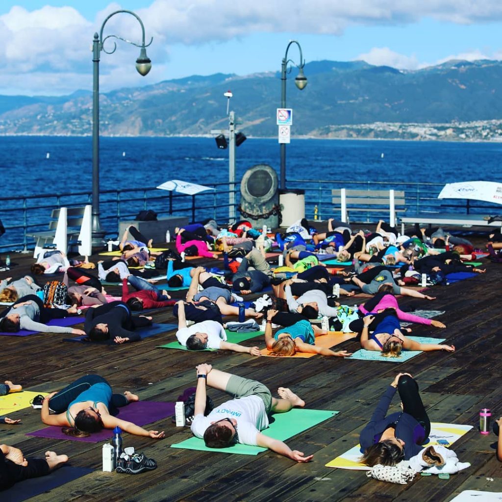 People doing yoga at the end of the Santa Monica Pier with the ocean and Santa Monica Mountains in the background.